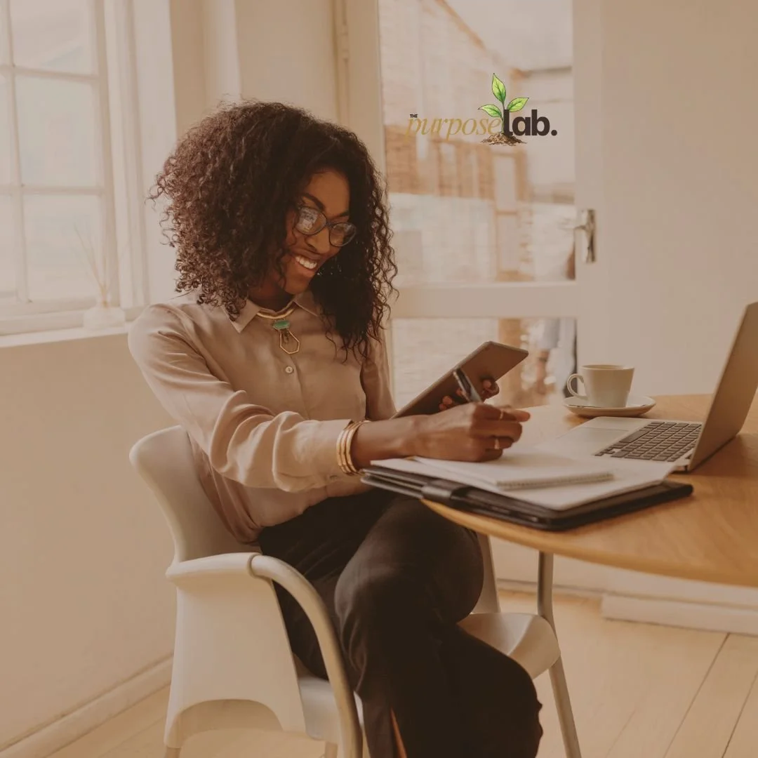 A woman with curly hair wearing glasses, a beige blouse, and jewelry sitting at a wooden table with a laptop, notebook, and a disposable coffee cup, smiling while looking at her phone inside an office space.