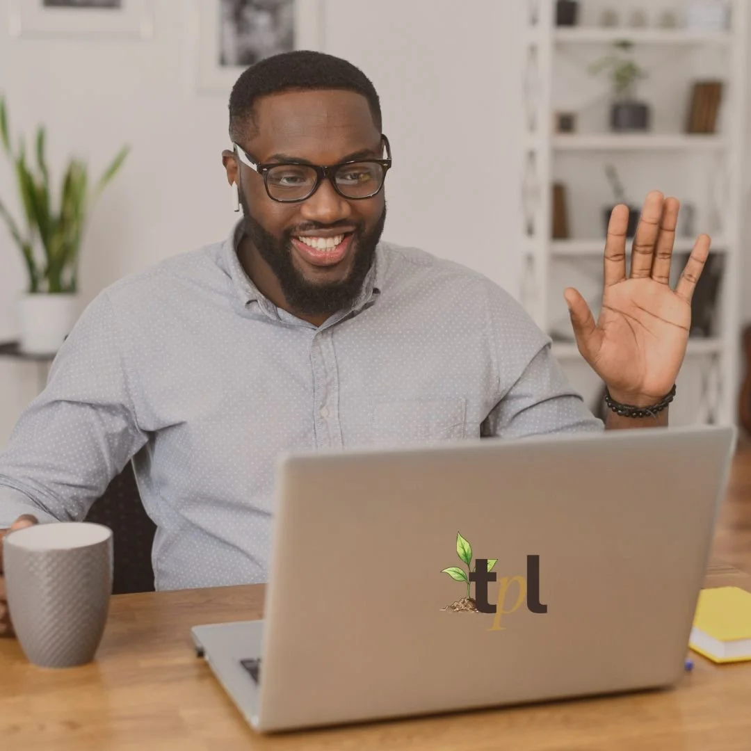 A smiling man with glasses and a beard sits at a desk, waving at a laptop with a logo featuring a plant and the letters 'tpl'. He has a coffee mug in front of him and is in a modern, organized room with shelves and a plant in the background.