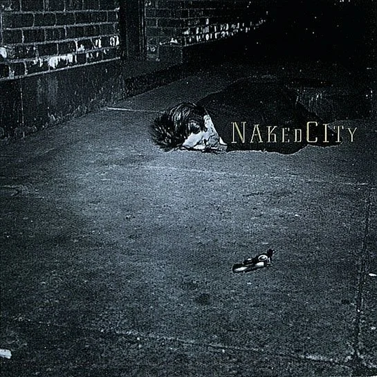 Person lying on the ground at night in an alley with a brick wall background, with the words Naked City illuminated nearby, and a gun on the ground.