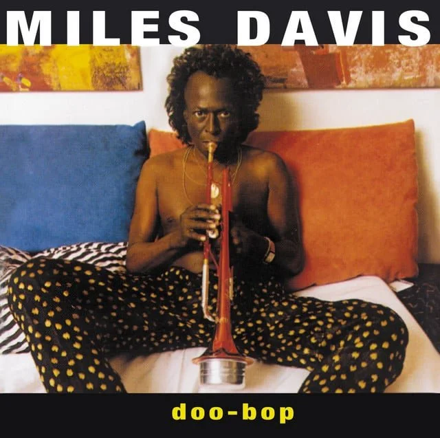 A man sitting on a bed with colorful pillows, playing a trombone, wearing black and yellow polka-dot pants, with the word "Miles Davis" at the top and "doo-bop" at the bottom.