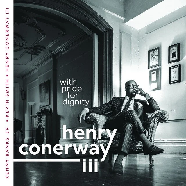 Black and white photo of a man sitting on a vintage sofa in a well-lit room with framed pictures on the wall. He is wearing a striped suit, glasses, and a pocket square, posing thoughtfully. Text on the image advertises an album titled "iii" by Henry