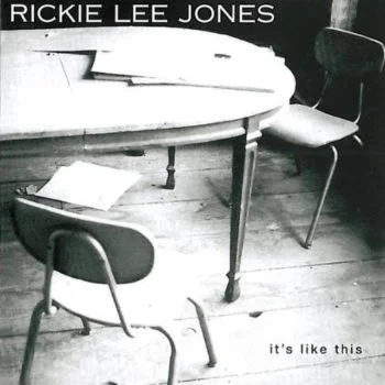 A black and white photo showing a round table with a few sheets of paper on it, two chairs around the table, one chair close-up and another pushed back, with a wooden floor. The text reads "RICKIE LEE JONES" at the top and "it's like this" at the bot