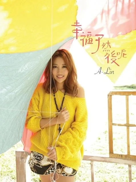 Young woman with long hair wearing a yellow sweater and patterned shorts, holding an umbrella and smiling, standing outdoors.