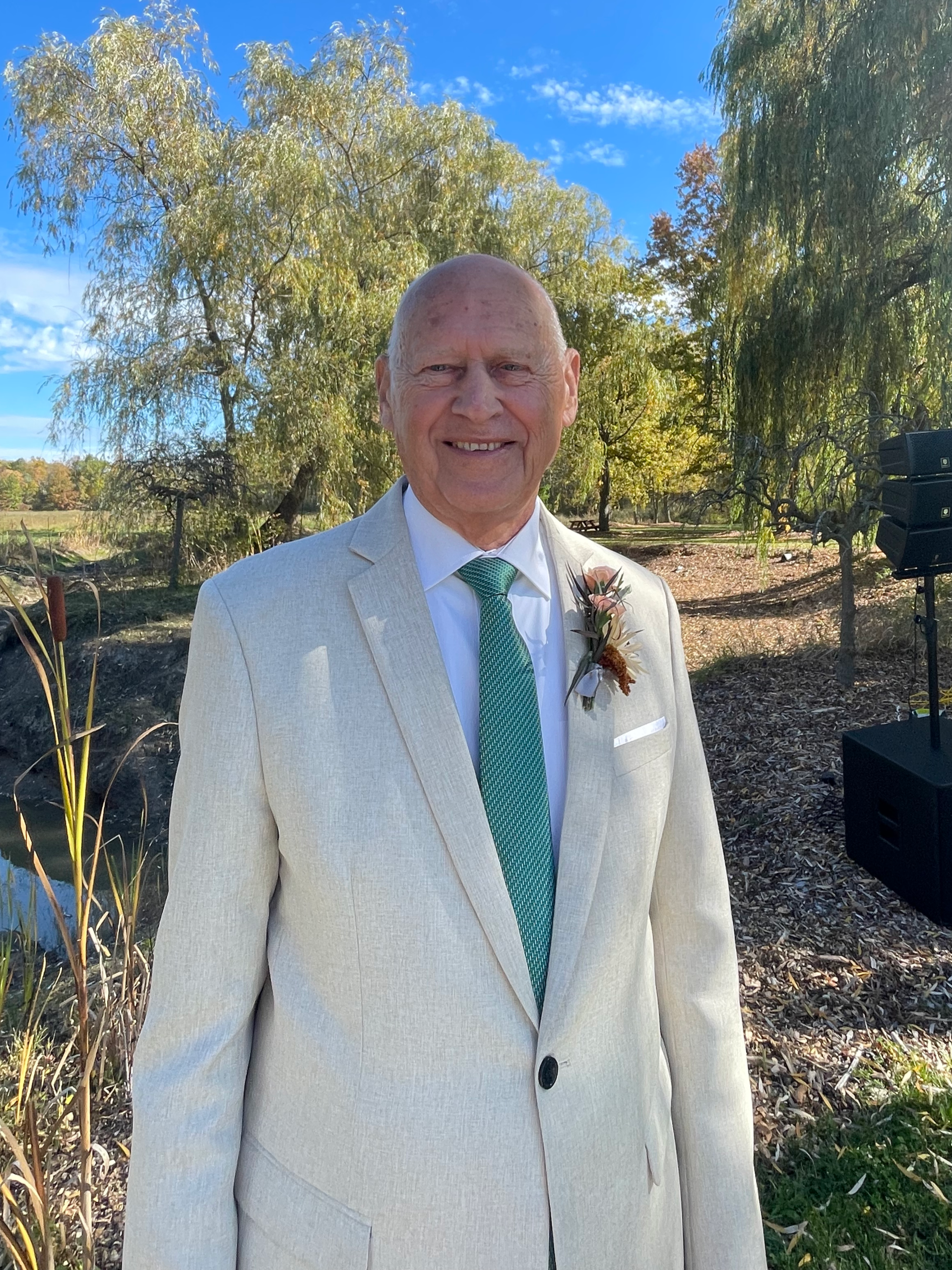 Photo of an elderly man wearing a light-colored suit, light blue shirt, and green tie, smiling outdoors with trees and a blue sky in the background.
