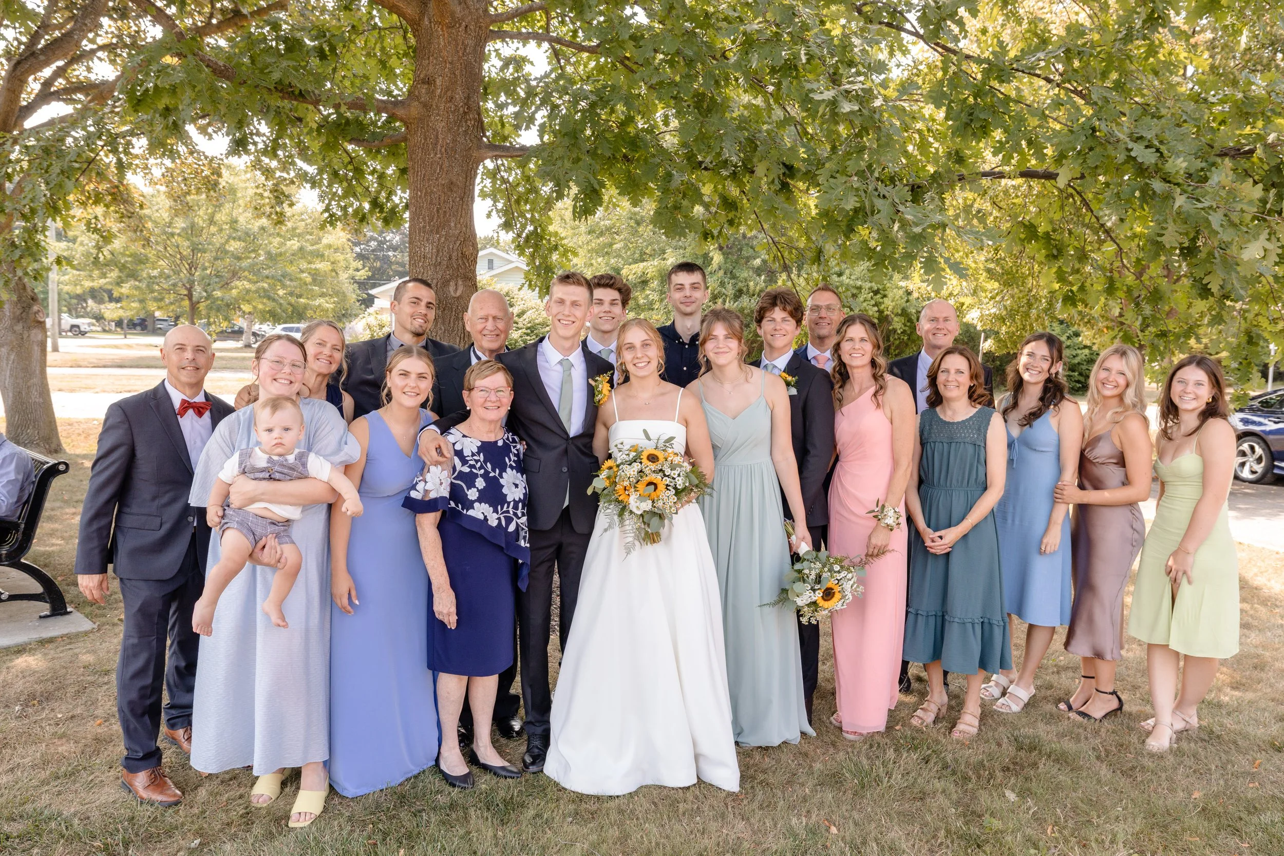 A large group of diverse people, including men and women of various ages, gather outdoors under a large tree for a wedding celebration. The bride and groom are at the center, with the bride holding a bouquet of sunflowers. Everyone is smiling and dre