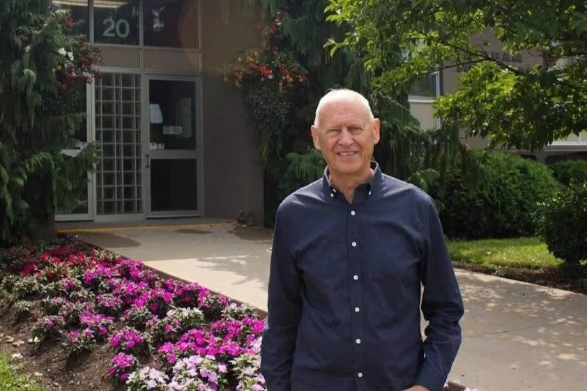An elderly man in a navy button-up shirt standing outside near a building with glass doors and windows, surrounded by colorful flowers and greenery.