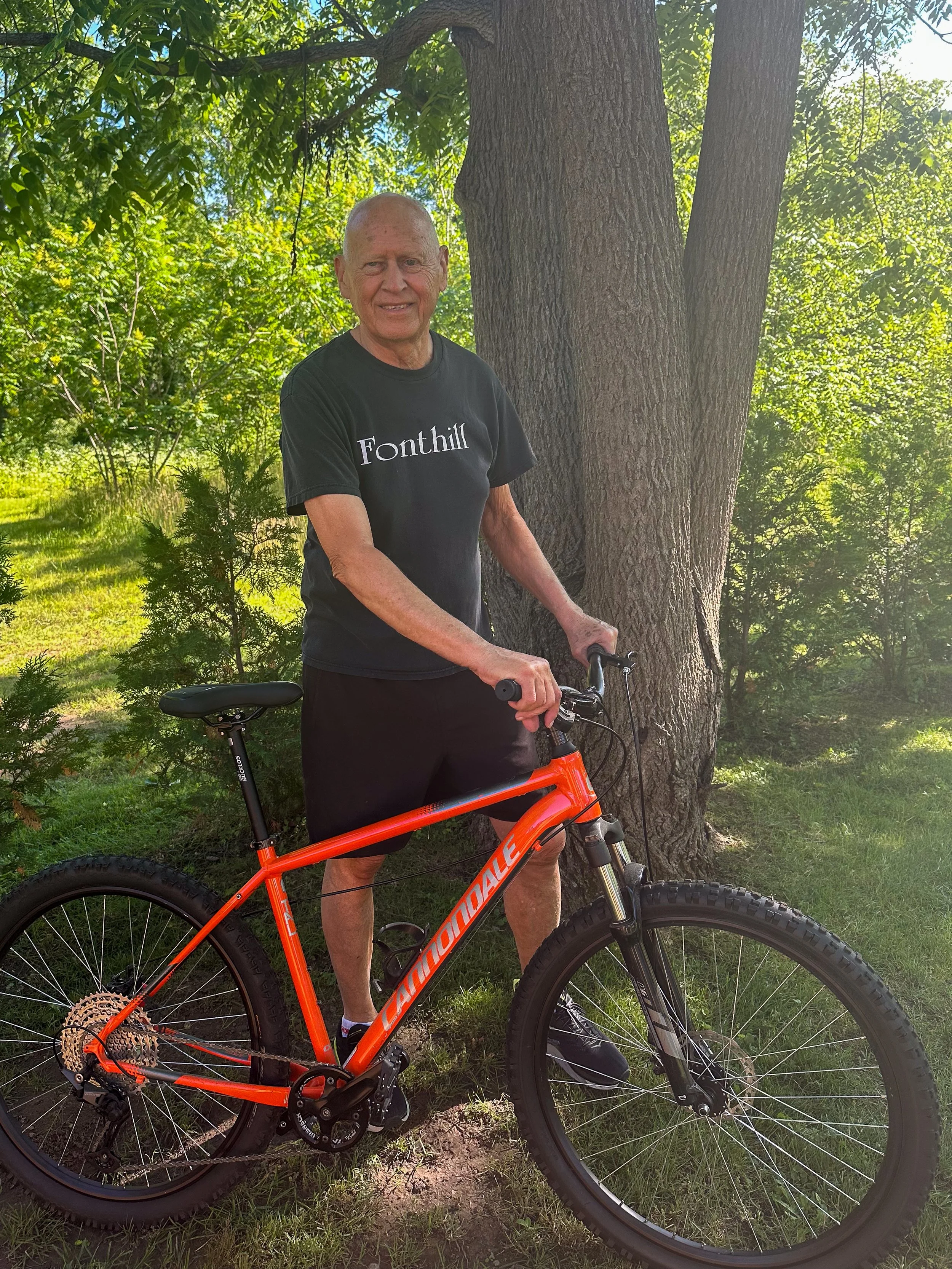 An elderly man with gray hair smiling while holding a bright orange mountain bike outdoors near a large tree with green leaves, sunlight filtering through the branches.