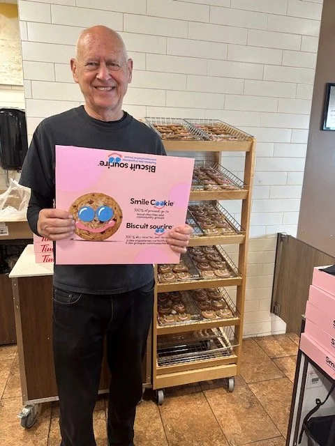 A man holding a pink sign featuring a smiling cookie character, standing in front of a rack of baked cookies in a store.
