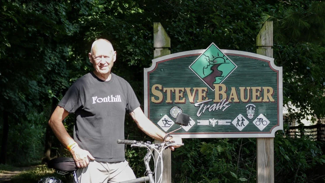 A man standing beside a bicycle next to a sign that reads 'Steve Bauer Trails' with icons representing outdoor activities, in a wooded area.