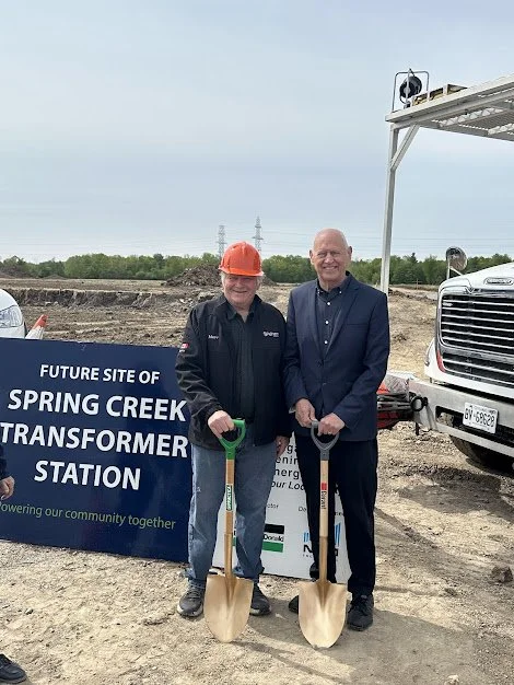 Two men standing with shovels at a groundbreaking event for the Spring Creek Transformer Station, with a sign behind them and construction site around.