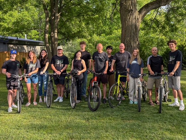 Group of ten people standing outdoors with bicycles, in front of a large tree and greenery.