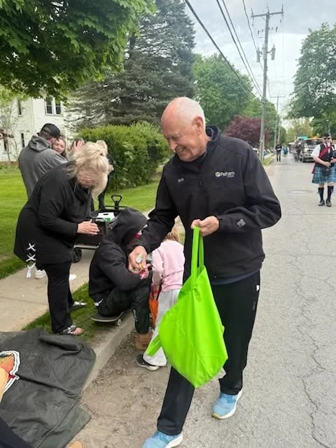 Man in a black jacket handing an item to a young girl on a sidewalk, with other children and people nearby, trees, and power lines in the background.