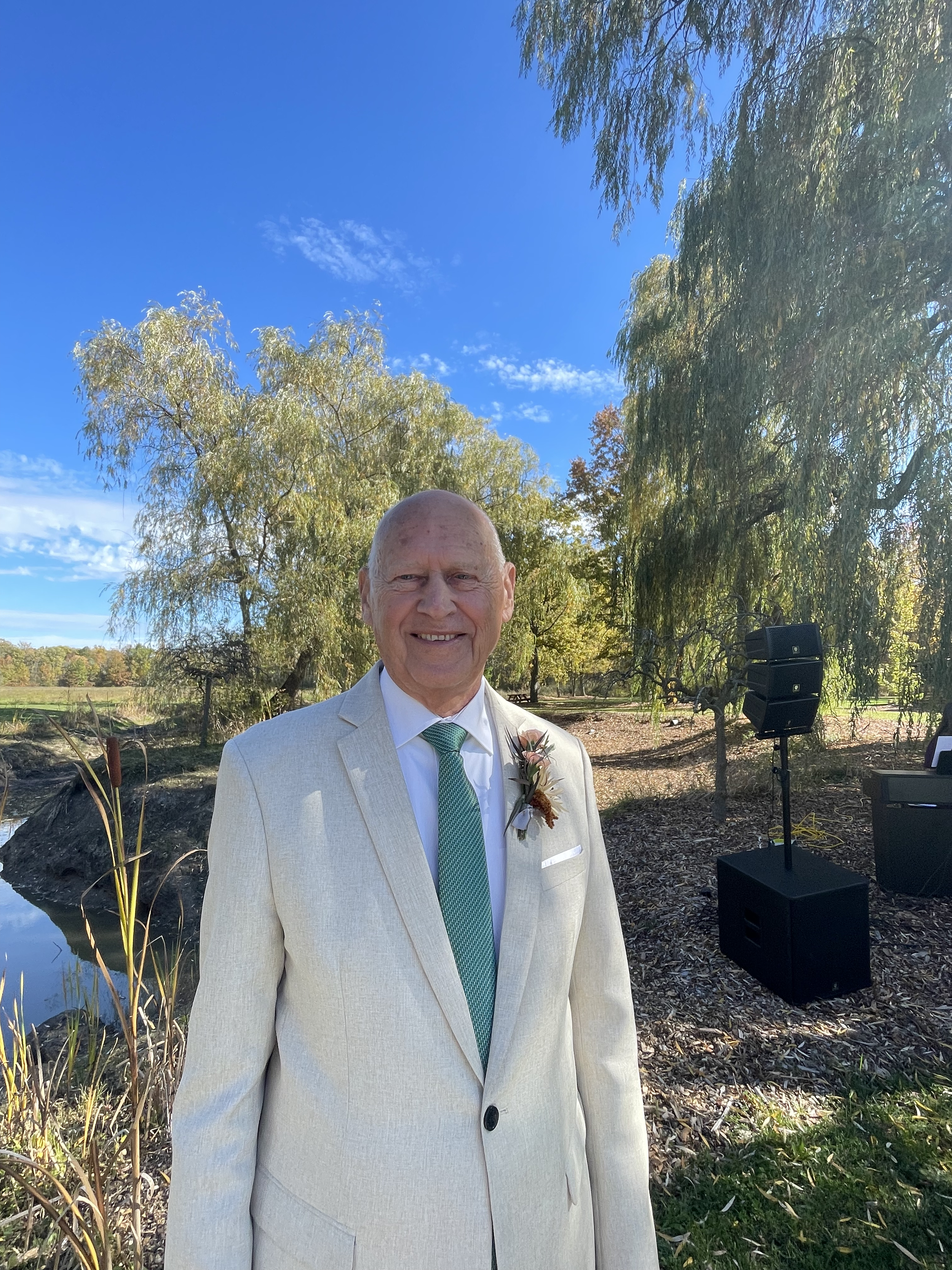 A man is smiling outdoors in a light-colored suit with a green tie and a floral boutonniere, standing in front of trees with a pond and speaker system nearby on a clear day.