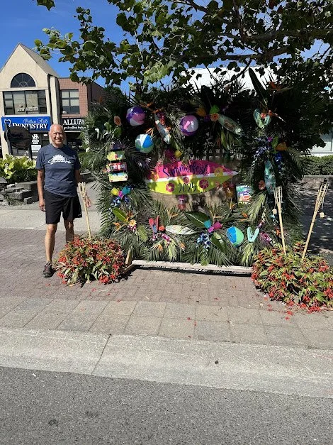 A man standing next to a decorated display with colorful eggs, butterflies, and a sign that reads 'Spring Fest 2022' on a sidewalk. There are buildings in the background and trees overhead.