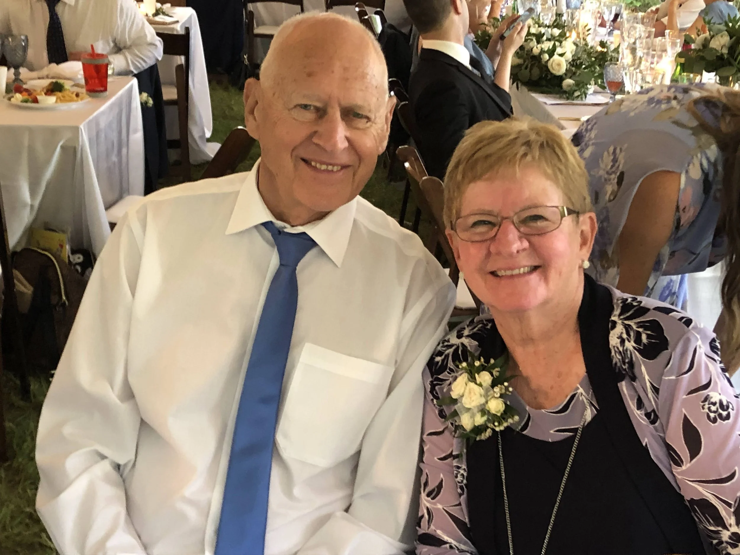 Smiling elderly man and woman at a formal event, sitting at a table with floral centerpieces, glasses, and floral arrangements in the background.