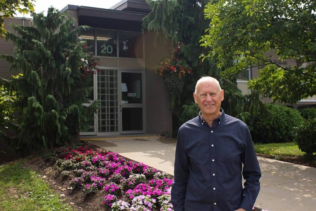 An elderly man with glasses and a smile, wearing a navy blue button-up shirt, standing outdoors in front of a building with a glass door, surrounded by green trees and a flower bed with pink and white flowers.