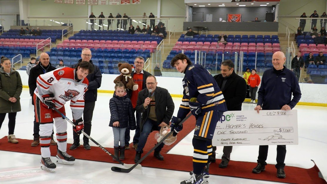 Group of people on an ice rink during a charity event, with children, hockey players, and a large check for $1,000.