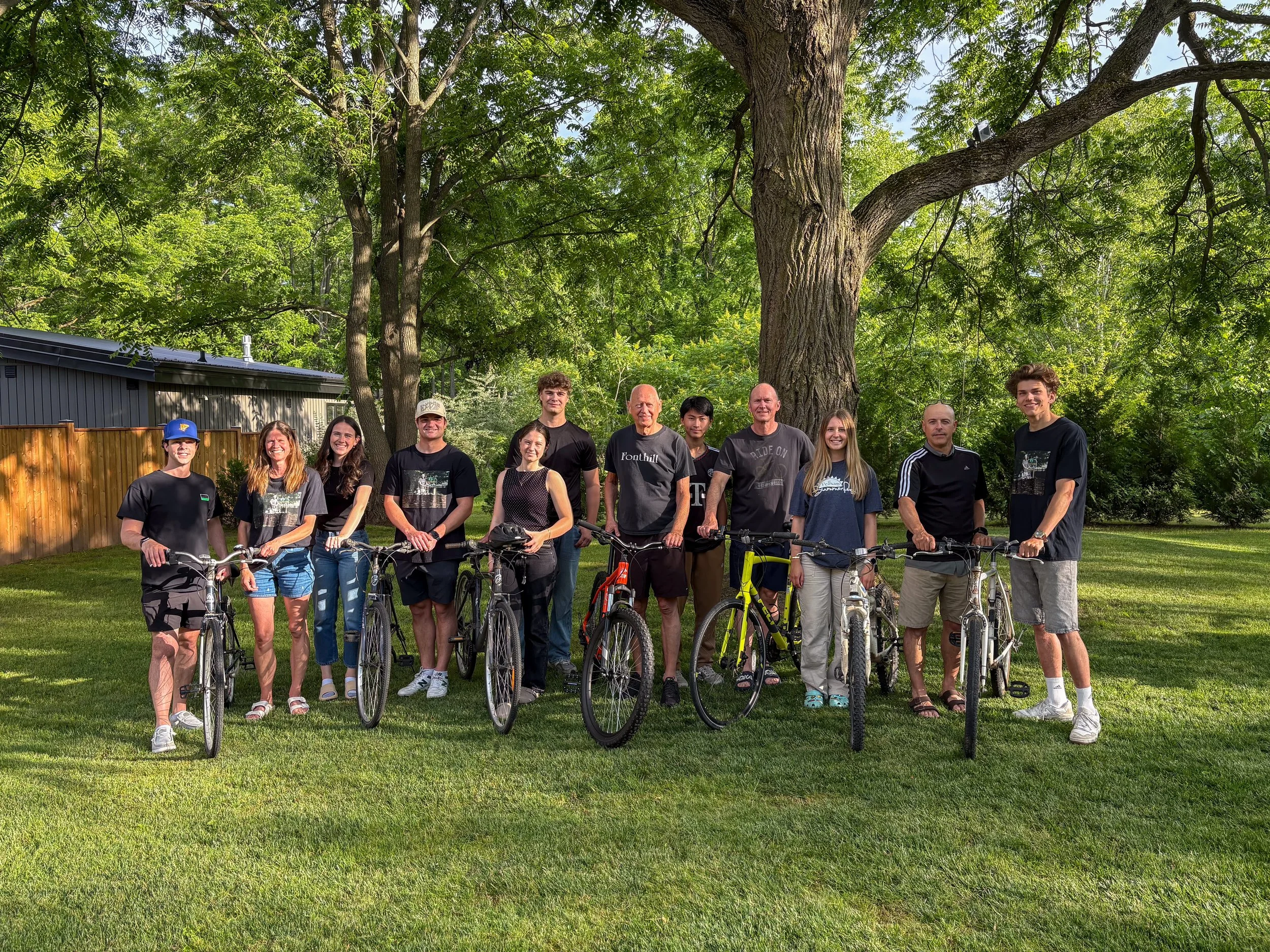 Group of people standing outdoors with their bicycles, smiling, under a large tree in a backyard with a wooden fence and a metal building.