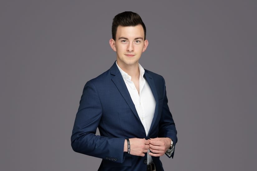A young man in a navy blue suit and white shirt standing against a gray background.