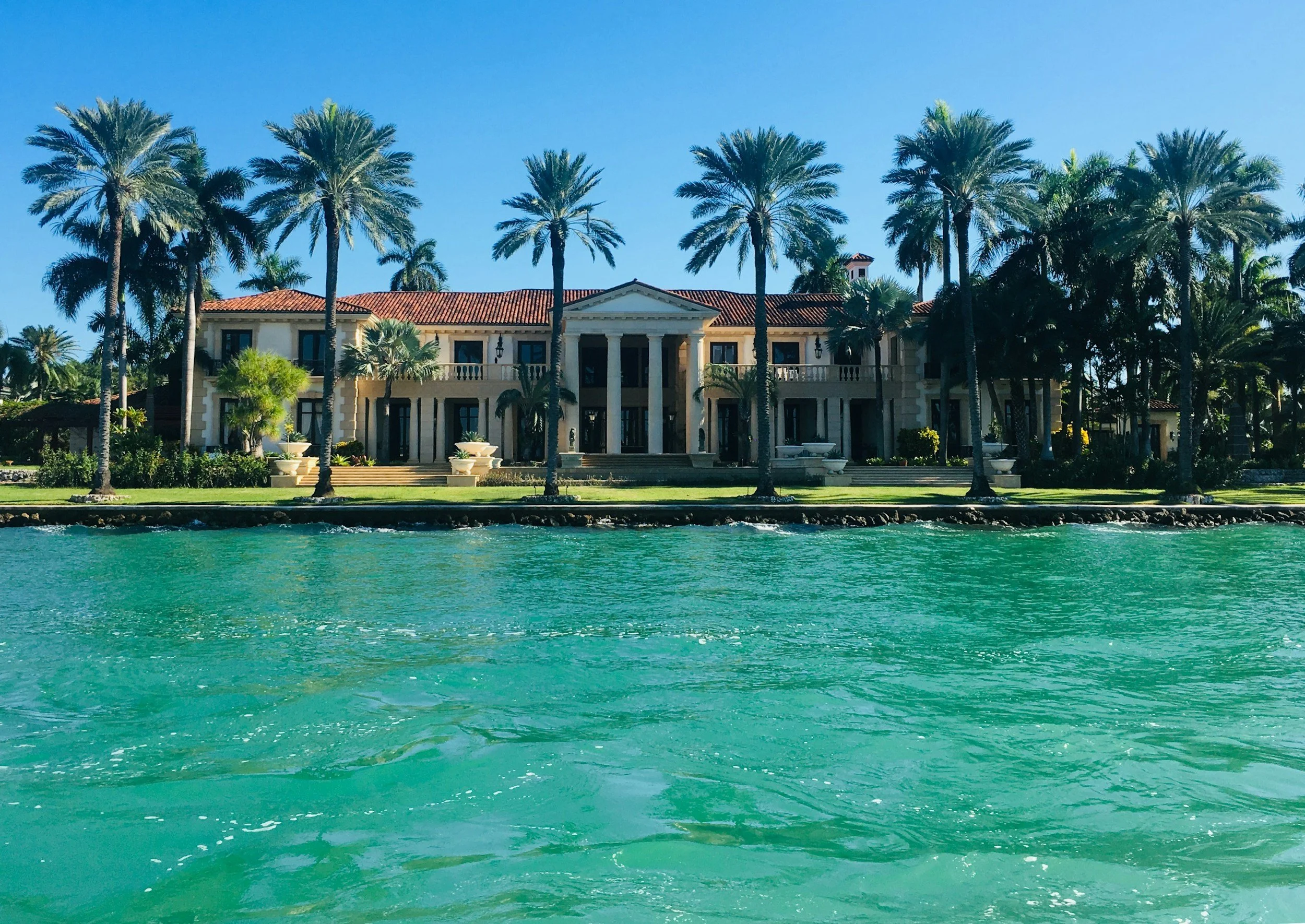 Luxury mansion with tall palm trees along a waterfront, clear blue sky, and turquoise water in the foreground.