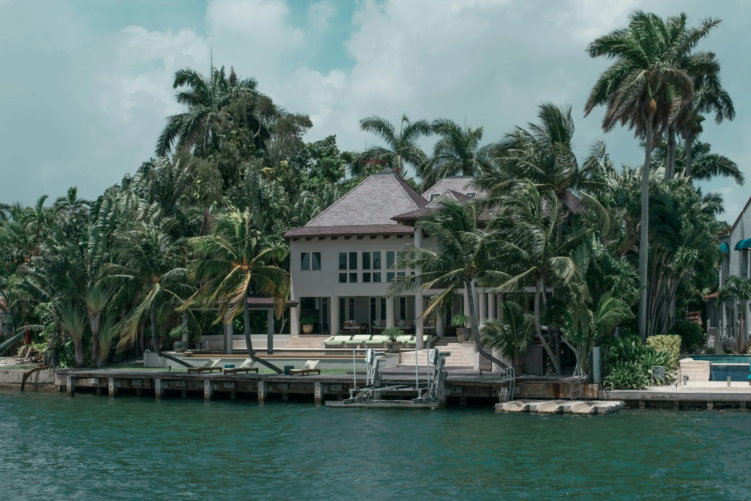 A luxurious waterfront house surrounded by tall palm trees with a swimming pool and lounge area in the backyard.