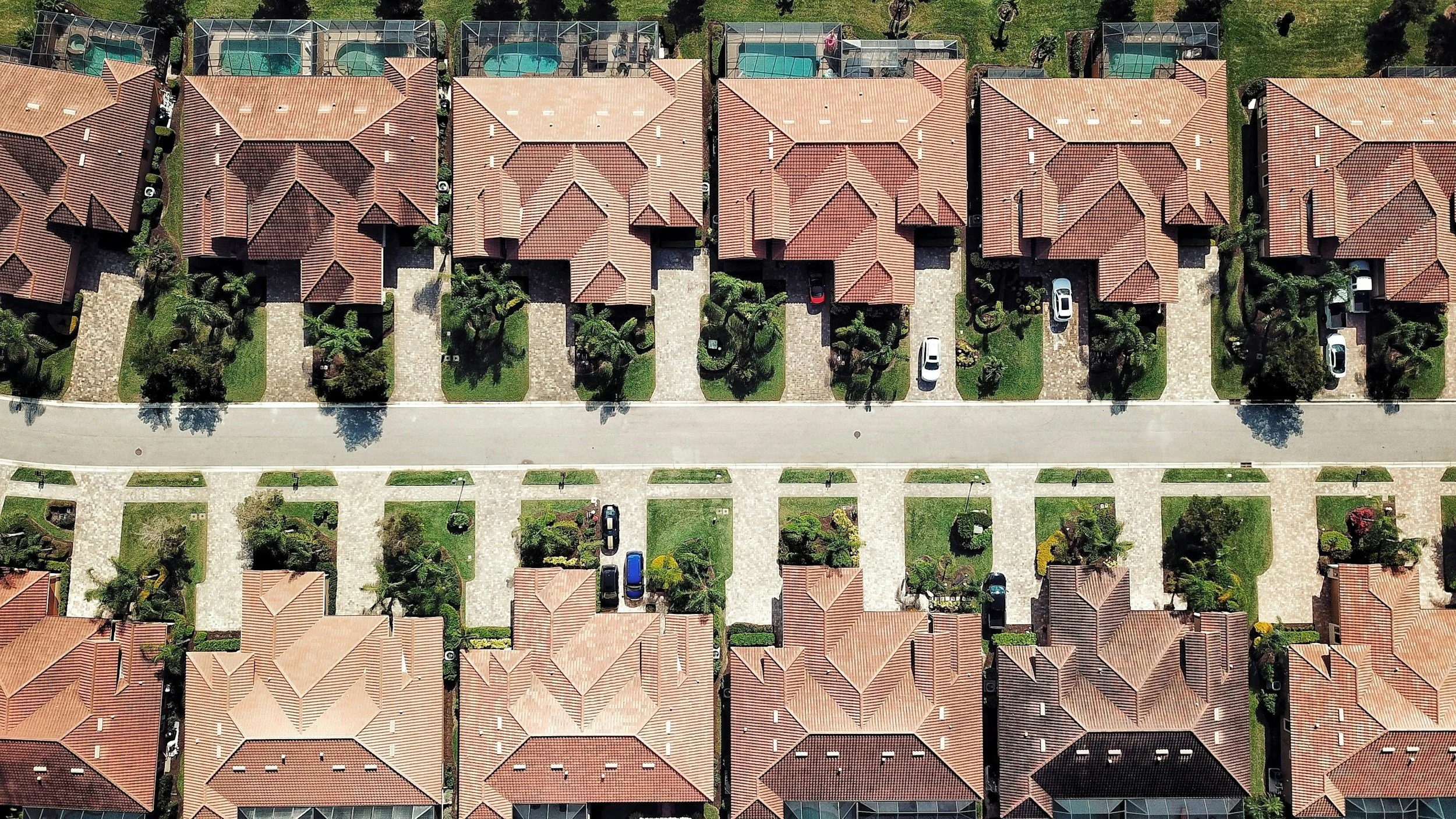 An aerial view of a row of houses with red tile roofs, each with a small front yard and driveway. Behind the houses, there are swimming pools enclosed in safety cages.