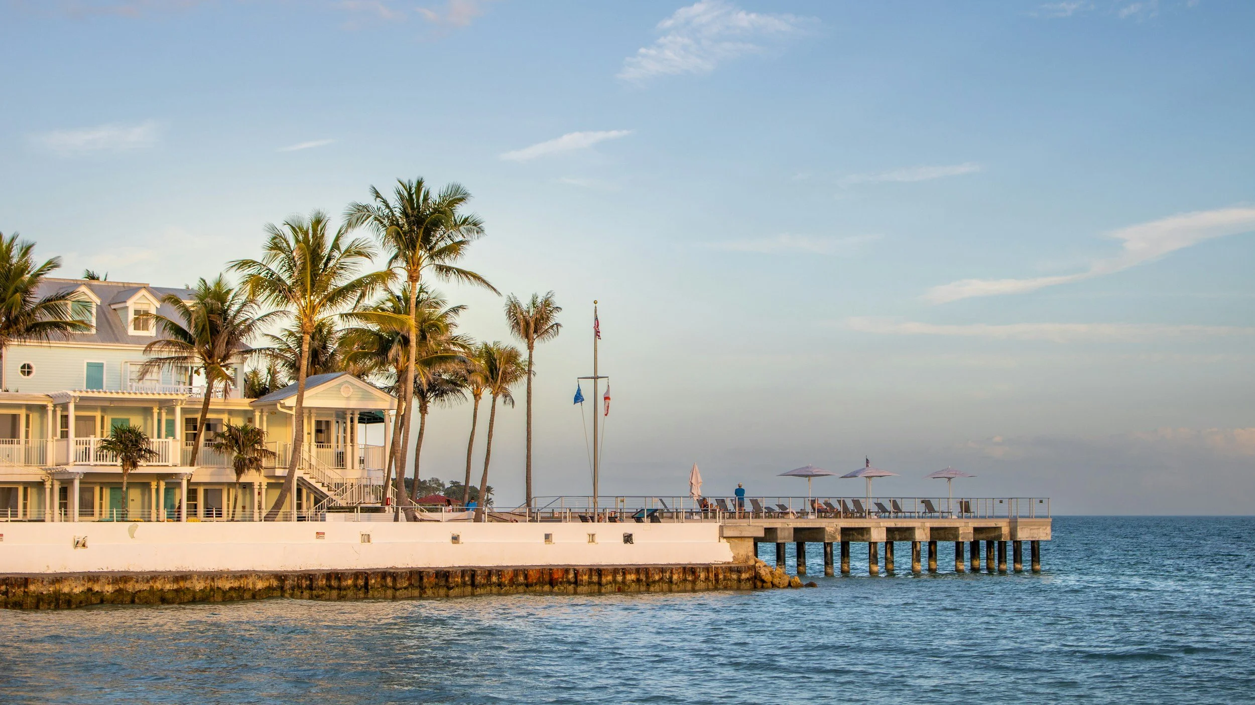 A seaside scene with palm trees, a multi-story building, umbrellas, and a pier extending into the water, under a partly cloudy sky during daytime.