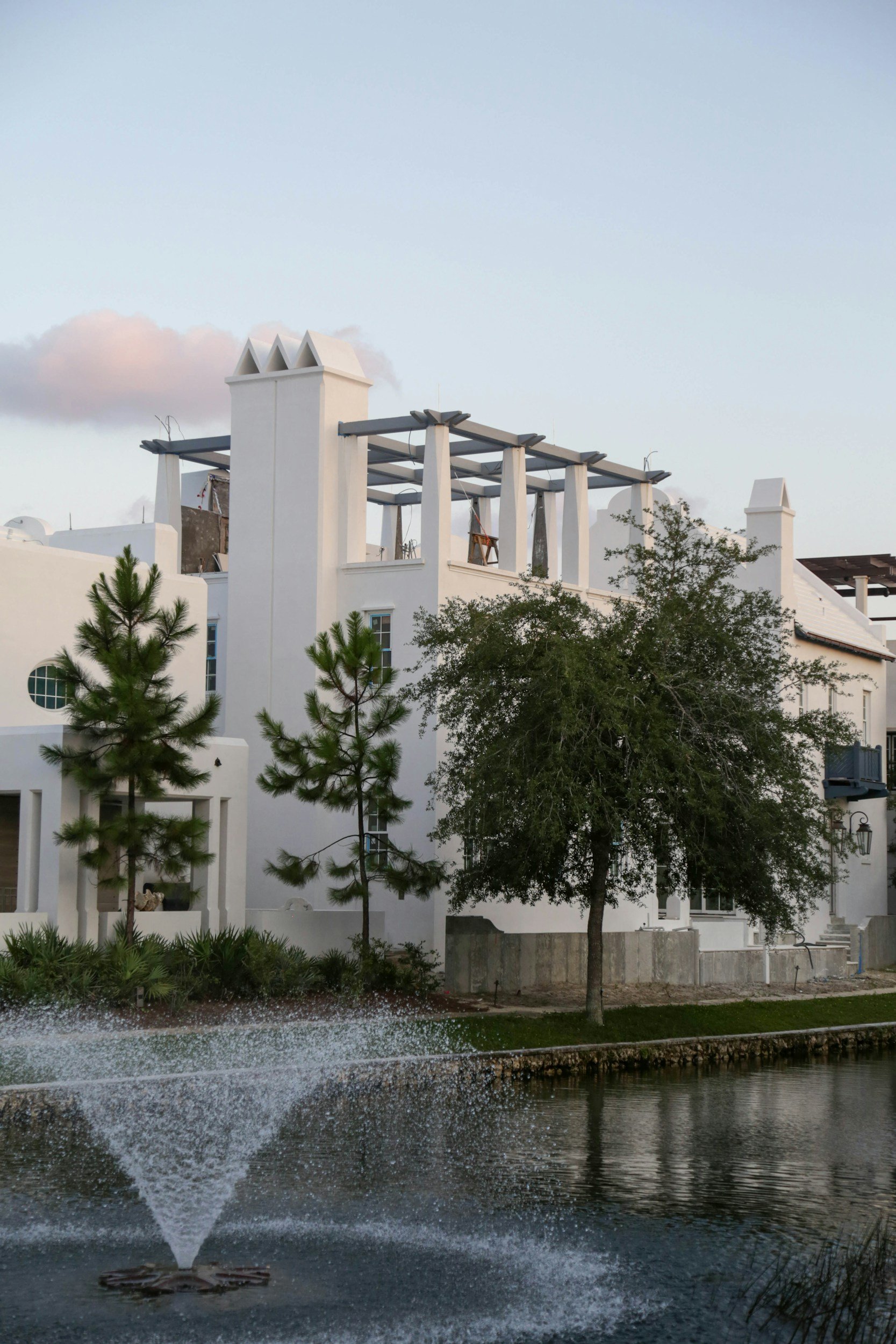 A row of white buildings with chimney-like structures on the roof, surrounded by trees and greenery, with a body of water in the foreground.