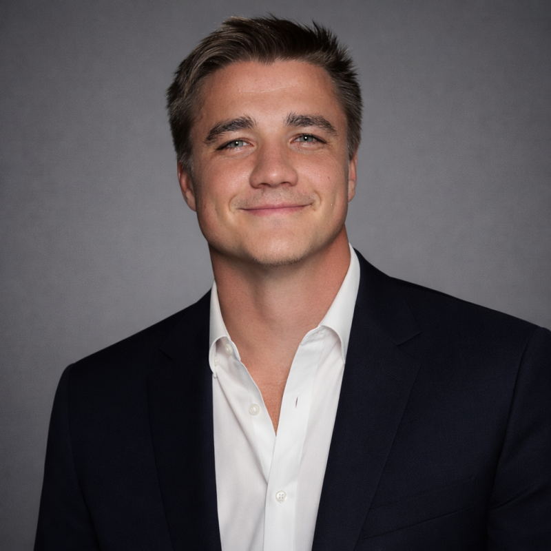 A young man with short, light brown hair, blue eyes, and a light complexion, wearing a dark suit jacket and a white shirt, smiling at the camera against a plain gray background.