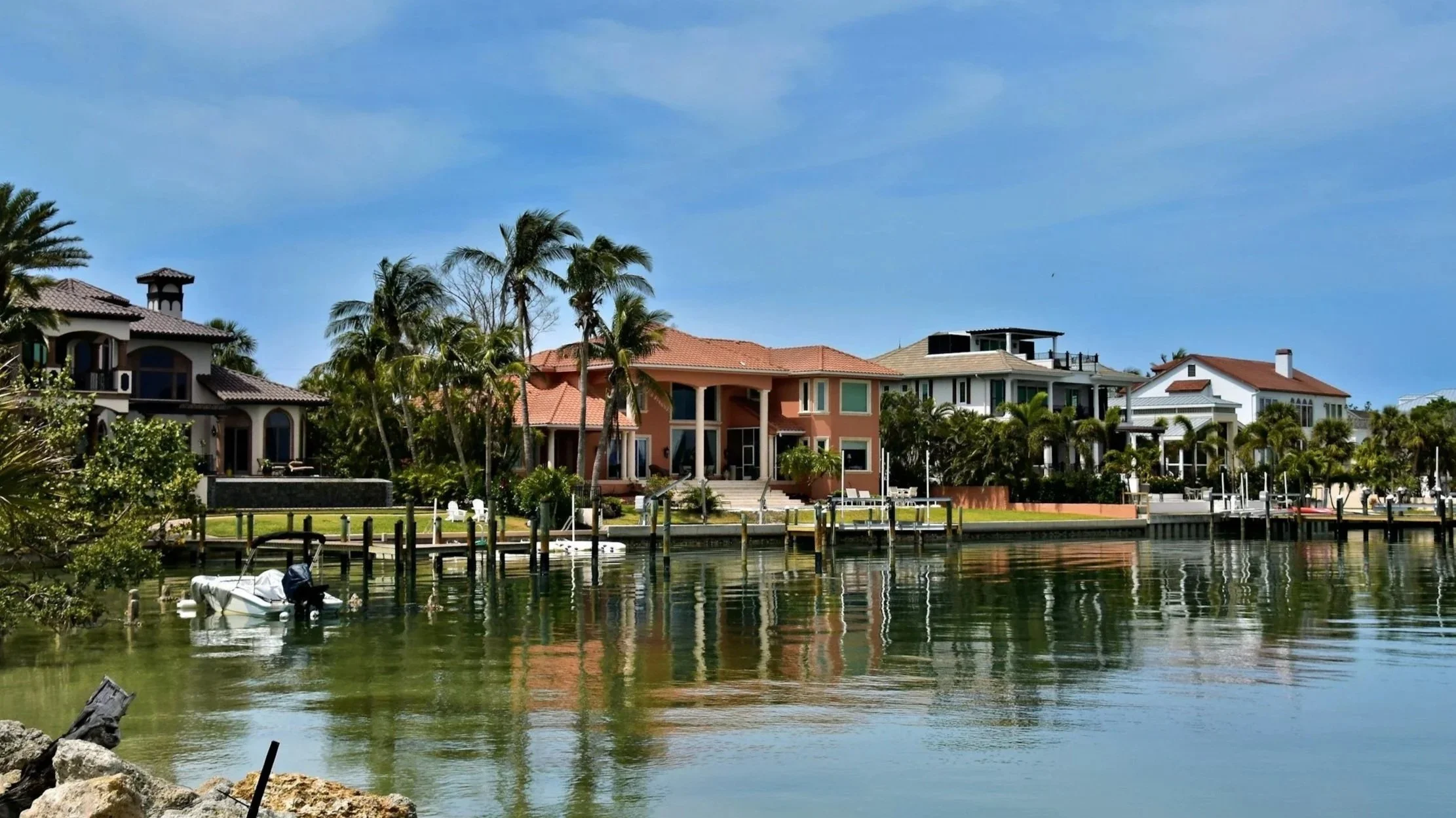 Luxury waterfront houses with palm trees, boats docked at a marina, and calm water reflecting the buildings and sky.