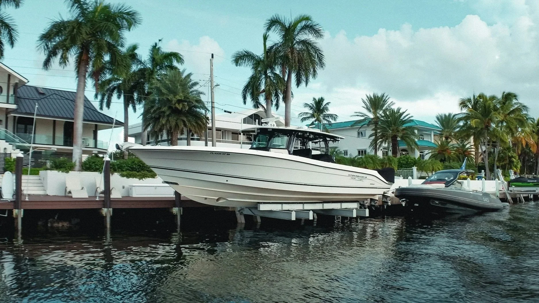 A marina with multiple boats docked along a waterway, including a large white motorboat in the center, with modern houses and palm trees in the background.