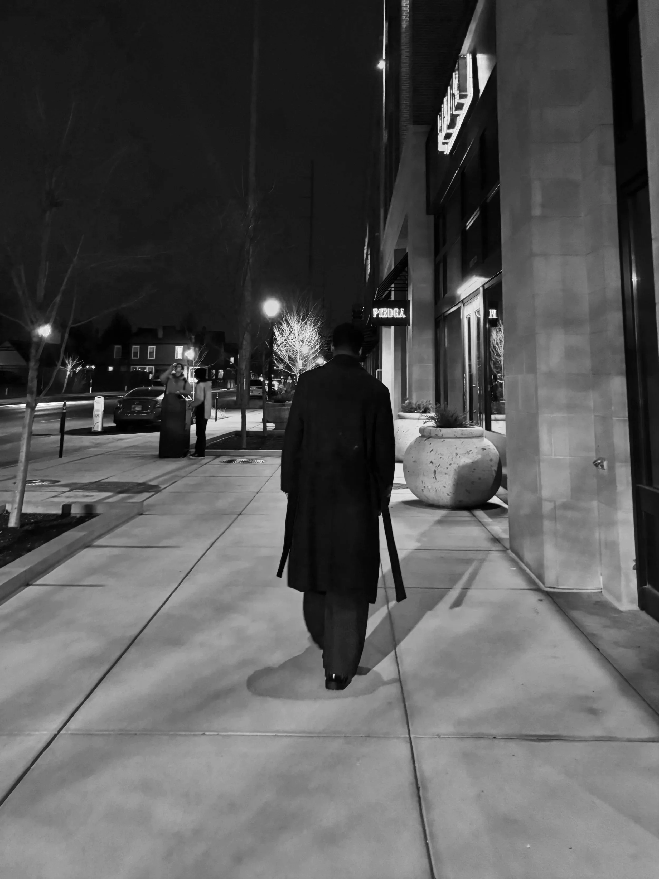 A man walking on a city sidewalk at night, carrying a cane, with a building on the right and other pedestrians in the background.