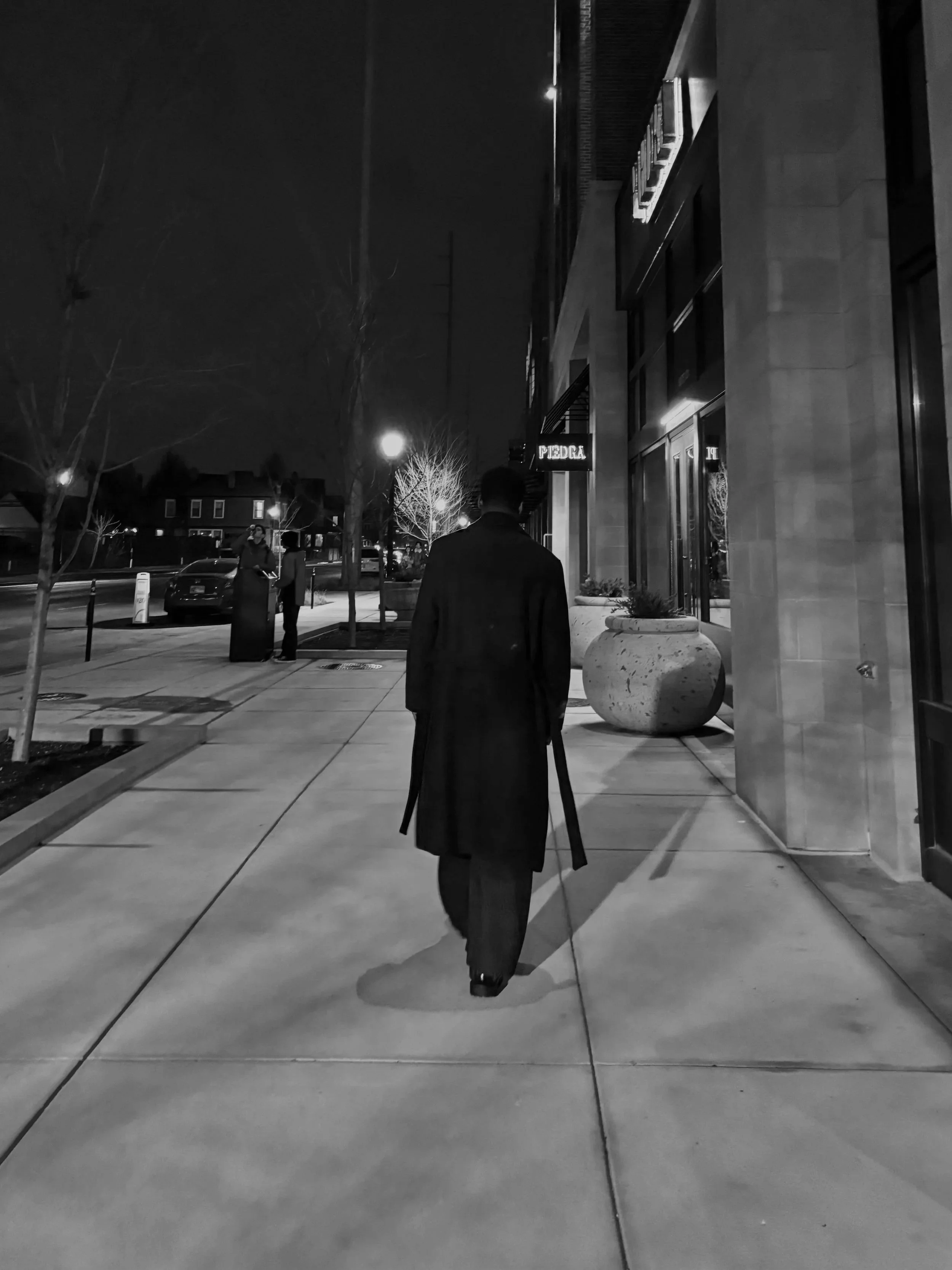 A man dressed in a long dark coat and hat walking with a cane on a city sidewalk at night, with buildings, streetlights, and trees in the background.
