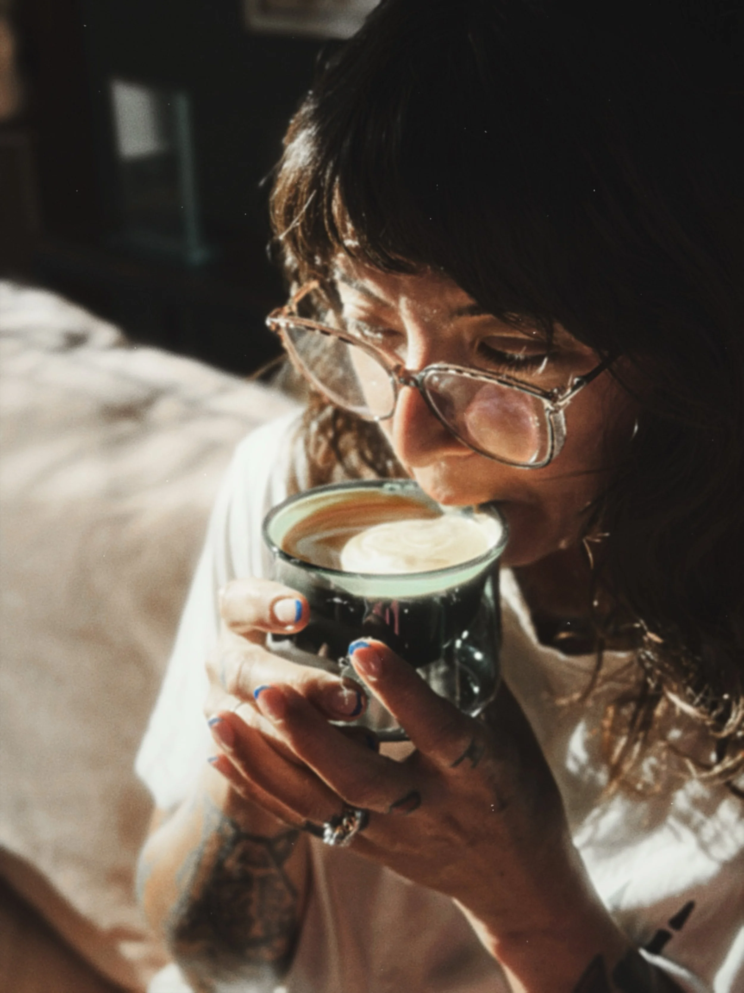 A person with dark hair, glasses, and tattoos on their arm is holding a coffee with latte art in a green glass mug and looking into it.