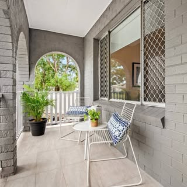 A balcony with a white table, two white chairs with blue and white cushions, a large potted plant, and a view of green trees through a large arched window.