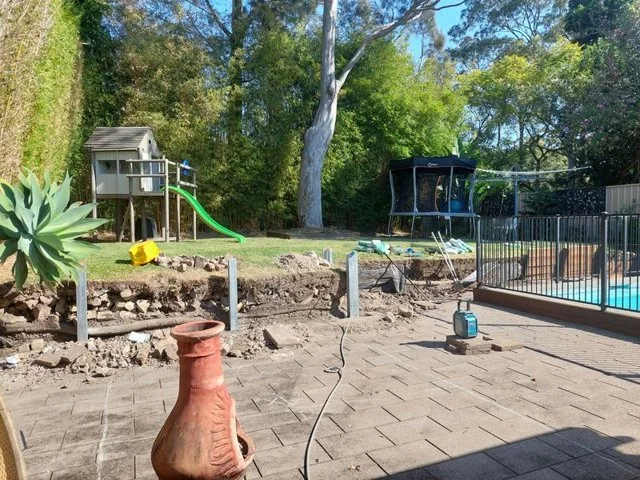 Backyard with a children's playhouse, slide, trampoline, and a swimming pool surrounded by a metal fence; construction or landscaping work in progress with tools and exposed ground.