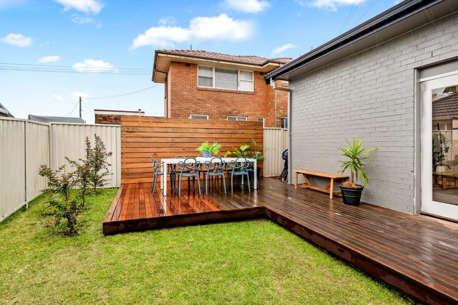 Backyard with wooden deck, metal table with six chairs, potted plants, and a white fence surrounding the yard. A gray brick wall is on the right side, and a small grassy area is in the foreground.