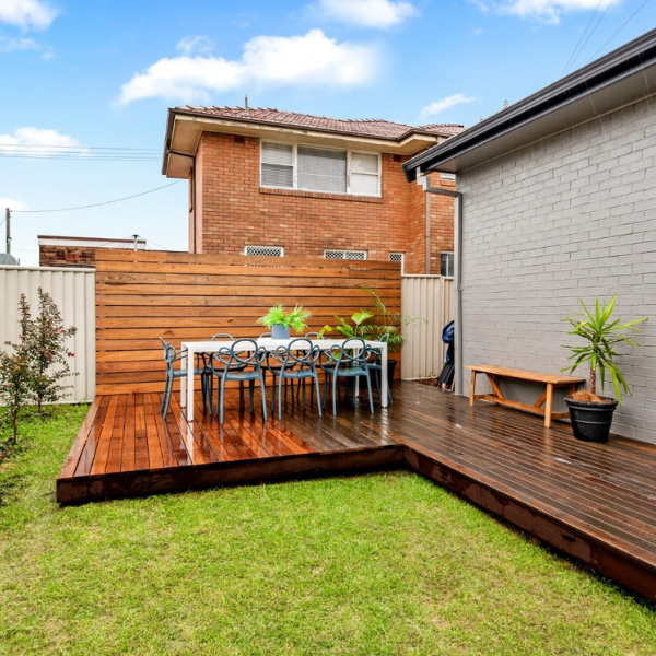 Backyard with a wooden deck, outdoor dining table with chairs, potted plants, and a white fence.