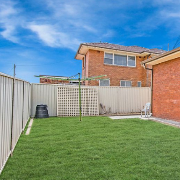 Backyard with green lawn, metal fence, clothesline, black compost bin, white plastic chair, and brick house in the background under a partly cloudy sky.