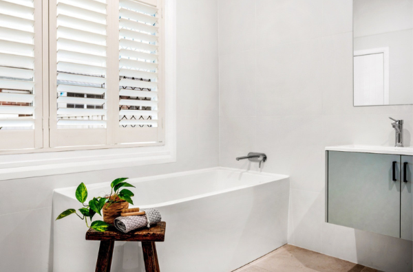 Modern bathroom with a white bathtub, a small wooden table with a potted plant and folded towels, white tiled walls, window with white shutters, and a wall-mounted sink with a mirror.