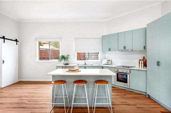 Modern kitchen with light blue cabinets, white island with three stools, wooden floors, and two windows with blinds.
