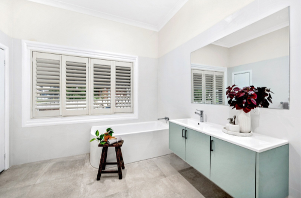 Bathroom with a large mirror, a white bathtub below a window with white shutters, a light blue cabinet with a sink, and a potted plant with dark red leaves on the countertop.