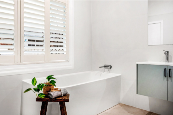 Bathroom with a white bathtub, window with white shutters, grey cabinet with black handles, and a small wooden stool with a potted plant and folded towels.