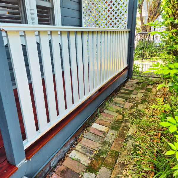 Close-up of a porch railing with a wooden top rail, painted white, beside a brick path and green bushes, with a gray house wall in the background.