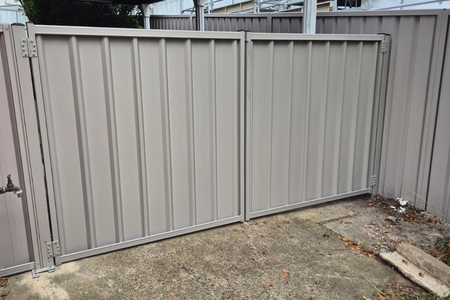 Metal backyard fence gate with a concrete ground and a small patch of dirt and leaves near the bottom right corner.