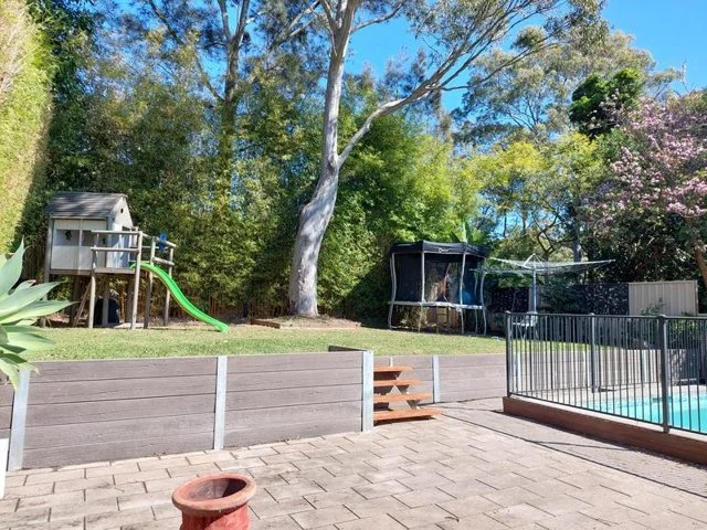 Backyard with a playhouse and green slide, trampoline, swimming pool with a metal fence, large trees, and a clear blue sky.