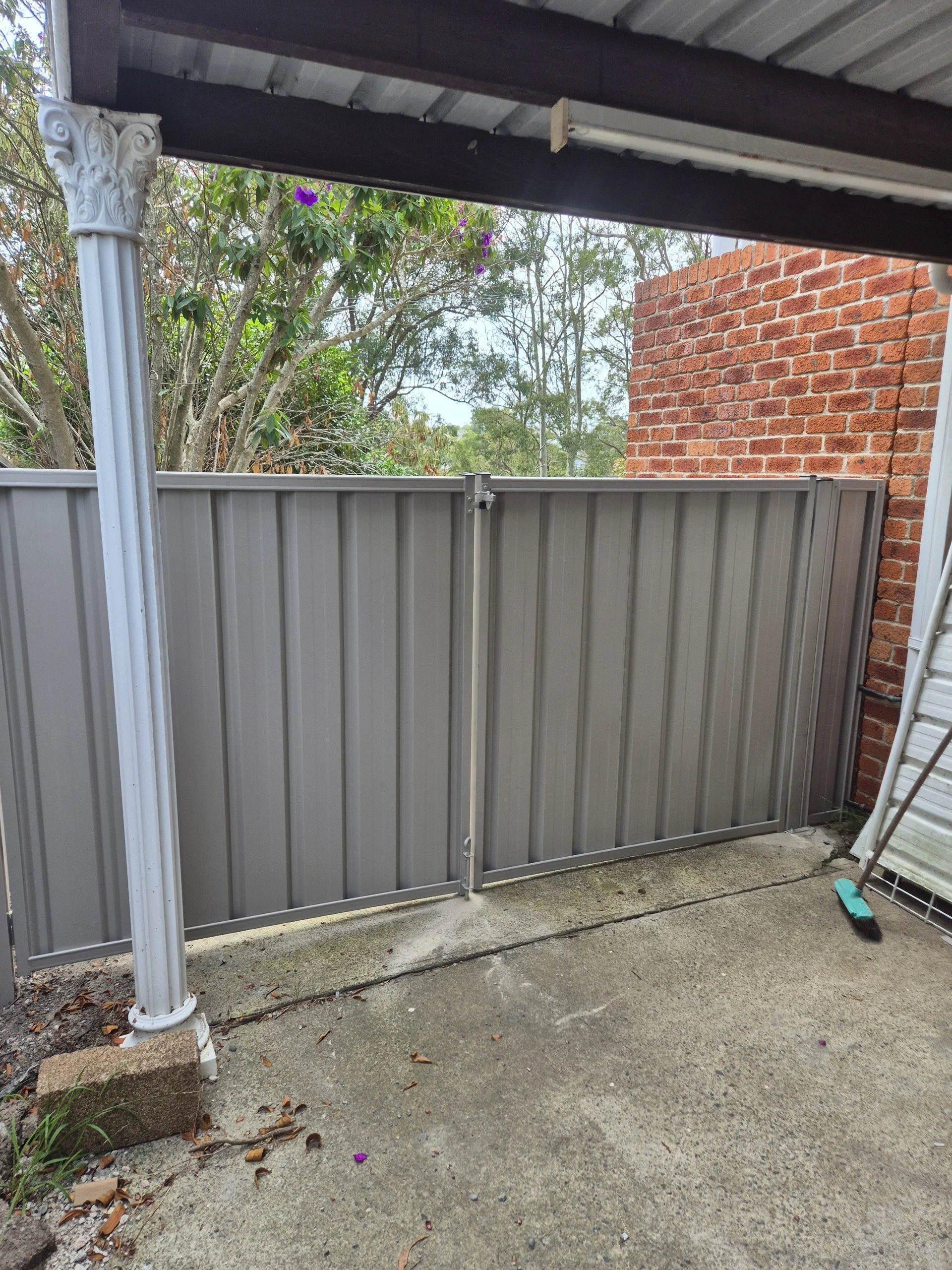 A gray metal gate with a latch, set in a concrete area outside a brick building, with a broom leaning against the wall on the right and some fallen leaves and debris on the ground.