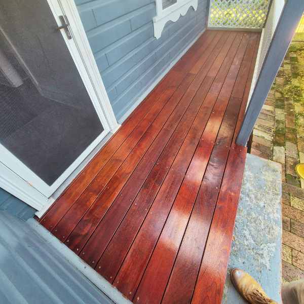 Wooden porch with reddish stain outside a blue house, a screen door, and a brick path. Part of a person's boot visible.