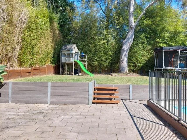 Backyard area with a small playground slide, steps leading up to it, a rock climbing wall, and a fenced swimming pool in the background, surrounded by trees.