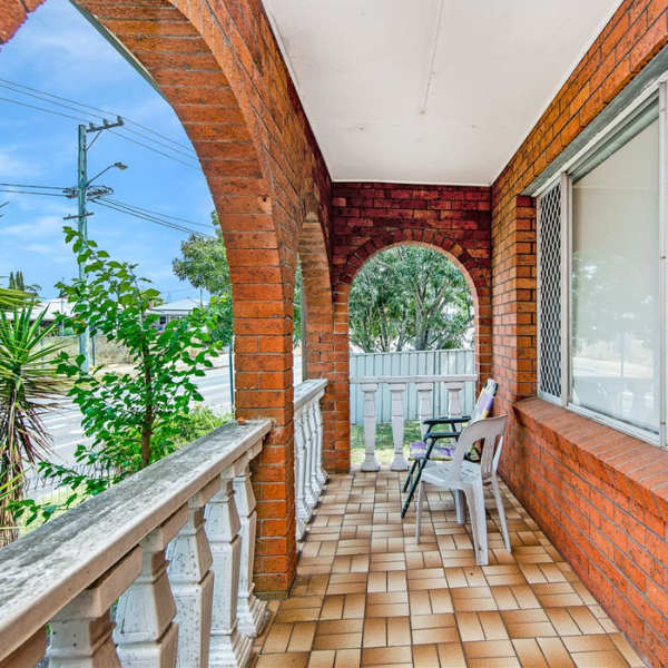 Covered front porch with brick walls, tiled floor, white railing, and two chairs facing outward, overlooking a tree and street.